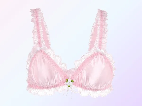 A baby pink satin bralette laid flat, ruffled lace along the straps and around the cups, a small white satin rose at the centre, on a soft pink and lavender gradient.